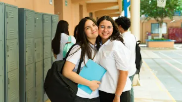 Portrait of two cheerful teenage girls wearing school uniform and embracing each other in a school hallway.