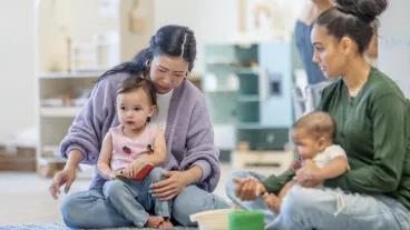 A warm interaction featuring mothers from different backgrounds engaging with their babies in a nurturing environment. Highlights creative play and early learning in a community-focused setting.