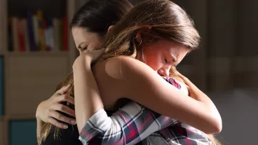 Side view of two sad good friends embracing in a bedroom in a house interior with a dark light in the background.