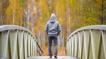 Young person in grey clothes slowly walking on bridge at natural park in colourful autumn day. Spending time alone in nature.