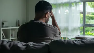 Young Asian man sitting on a sofa at home, looking sad and exhausted, struggling with depression and mental health issues.