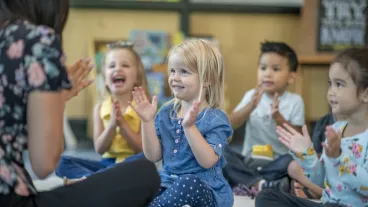 A small group of multi-ethnic preschool children sit on the floor in front of their teacher as she teaches them a song. They are each dressed casually and sitting with their legs crossed as they clap and sign along.