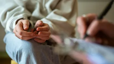 Close-up image of a teenage girl in casual clothes nervously fidgeting during a mental health therapy session with a professional taking notes.