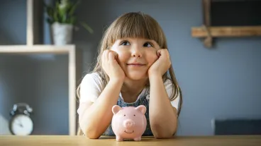 A young girl sits at a table in front of a piggy bank with her hands on her cheeks.