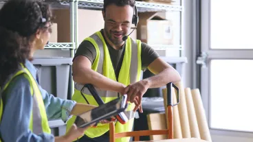 2 workers in a distribution warehouse wearing high vis clothing using a digital tablet.
