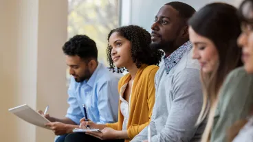 A group of business people sit in a row in a training class. They look at an unseen speaker as they concentrate on the lecture.