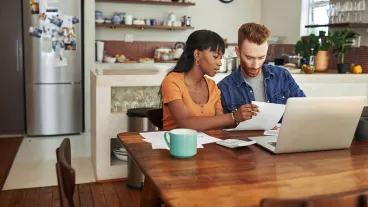 A couple reviewing paperwork and looking through the laptop on the kitchen table A couple reviewing paperwork and looking through the laptop on the kitchen table
