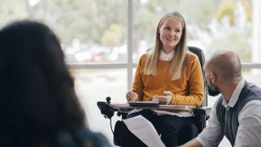A woman looks happy and confident as she leads a group discussion at her place of work. She is a wheelchair user and has Muscular Dystrophy.