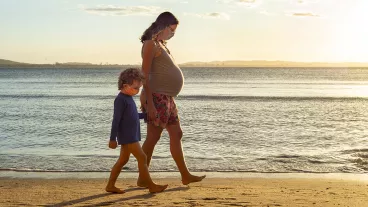 Pregnant mother exercising at the beach, holding her toddler's son hand by the sea, during sunset.