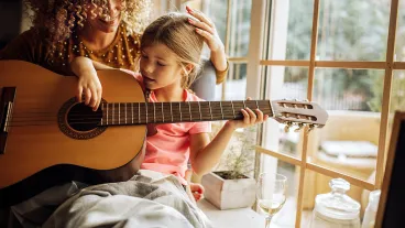 Adult woman at home during quarantine with her young daughter who is playing a guitar