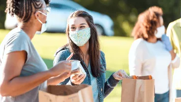 Two cheerful female friends fill paper bags with food donations as they volunteer during an outdoor food drive. They are wearing protective face masks as they are volunteering during the COVID-19 pandemic.