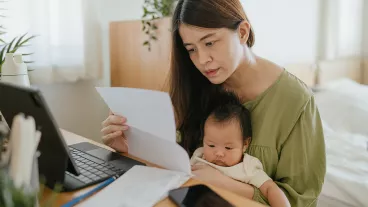 Mother sitting with baby in her lap, using laptop to check finances at home.
