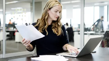 Businesswoman with long blond hair holding document and using laptop in modern office