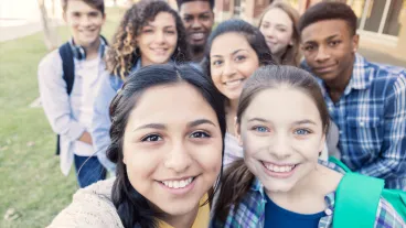 Diverse group of eight high school students are smiling and looking at the camera. Teenagers are students at public high school, and are wearing backpacks or holding school books.