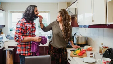 Mature woman is cooking a curry in the kitchen of her home. She is feeding her son a piece of sliced pepper as he does the dishes.