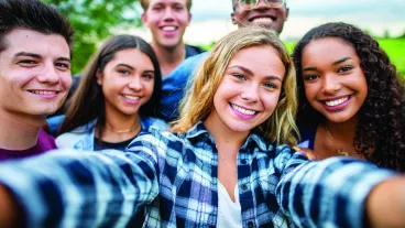 A multi-ethnic group of high school students are outdoors on a summer day. One girl is taking a selfie with the group.