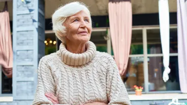 Waist-up portrait of pensive senior woman with crossed arms standing outdoors and looking away, facade of modern cottage on background