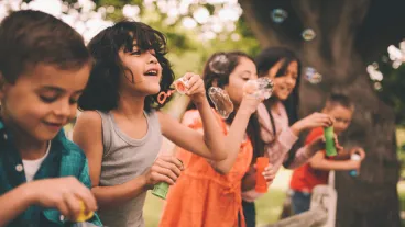 Long haired boy laughing and having fun with his friends standing on a wooden fence in a summer park blowing bubbles, with a vintage develop