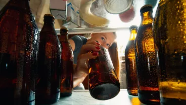 Young woman takes cold refreshing beer from out the fridge, inside view from fridge of hand holding the bottle.