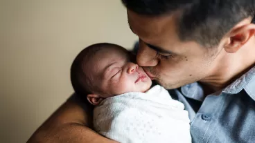A father gently kisses his newborn daughter on the cheek. He is holding her cradled in his arms as she is swaddled up comfortably for her nap.