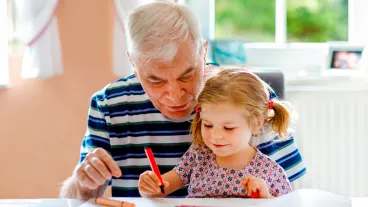 A senior man and his granddaughter are sitting on at a table colouring on a piece of paper.