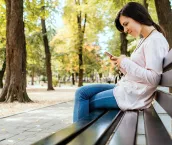 Young woman sitting on the bench in public park smiling and checking social media or messages on smartphone.