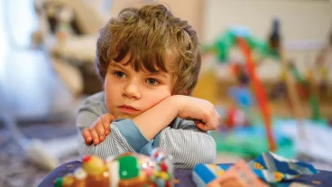 Image of a young boy sitting at a table with some colourful toys and he is resting his head on his arms. 